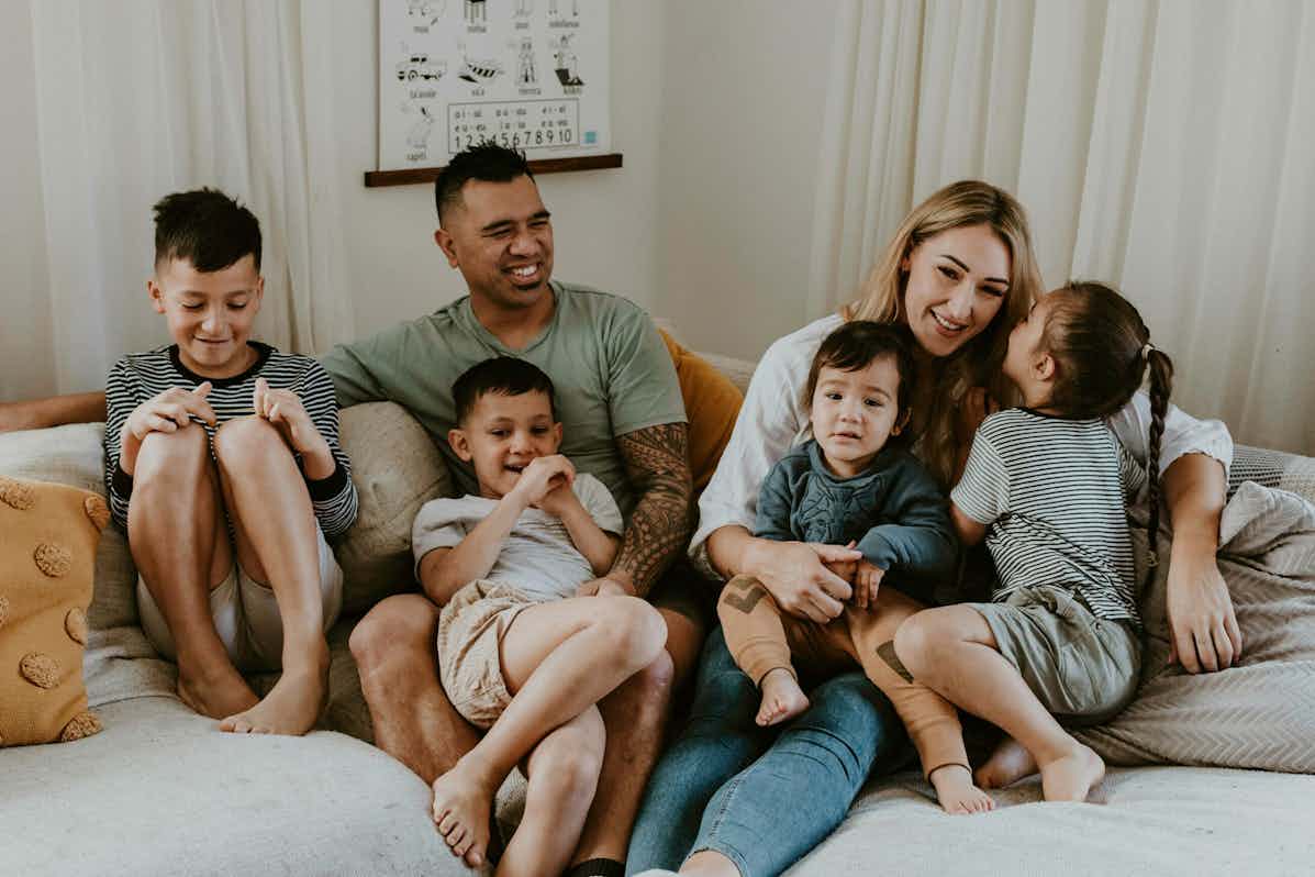 New Zealand family of relaxing on couch. Four children under the age of 8 years with mum and dad. Young daughter whispering on mums ear.