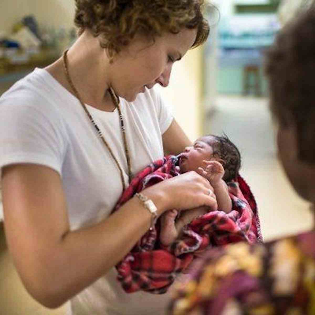 Woman wearing white top holding baby wrapped in red blanket.